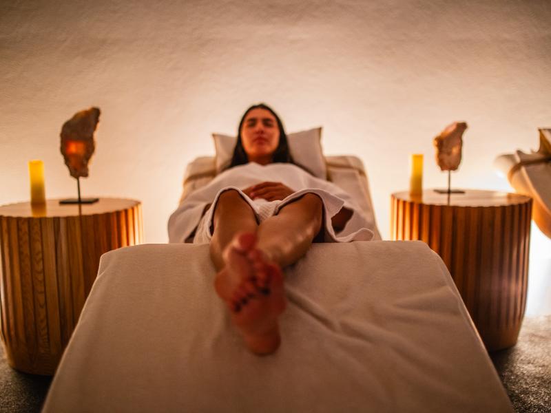 Osoba u belom ogrtaču leži na stolu za masažu u slabo osvetljenoj spa sobi sa ukrasnim bočnim stolovima i mekim osvetljenjem.