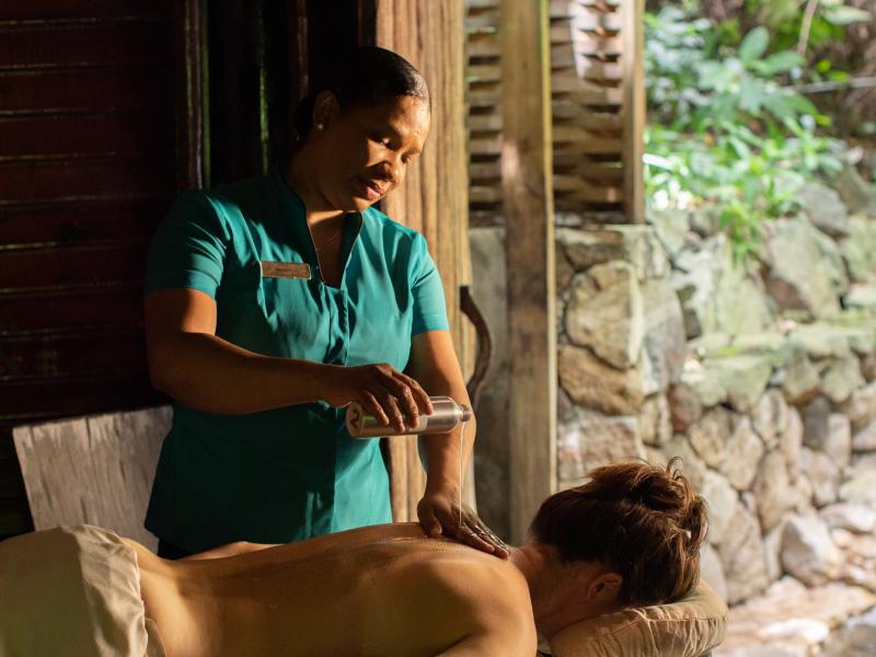 A massage therapist pours oil onto a client's back in an outdoor spa setting.