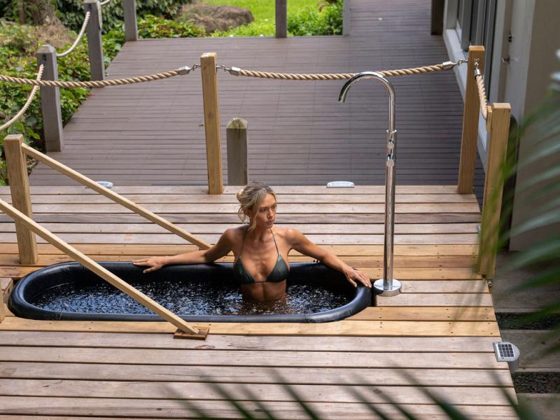 A woman in a bikini relaxes in an outdoor hot tub on a wooden deck surrounded by lush greenery.