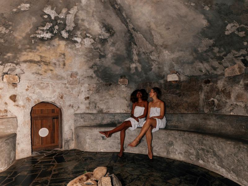 Two women in white towels sit on a stone bench in a domed, rustic room with a wooden door.