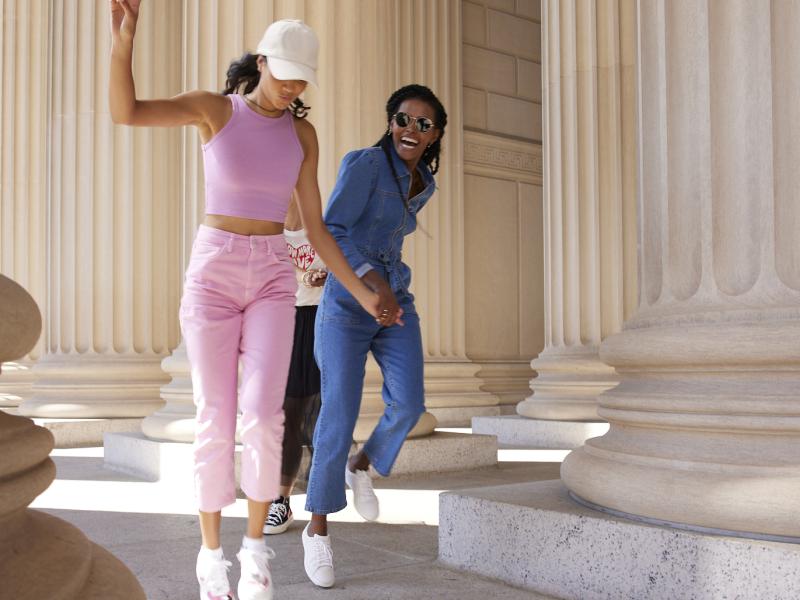 Deux jeunes femmes vêtues de tenues tendance se promènent en s'amusant entre de grandes colonnes classiques ; l'une d'elles fait le signe de la paix.
