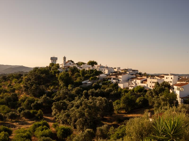 Elevated wide view of a small Portuguese village with white houses, surrounded by green trees and hills under a warm, hazy sky.
