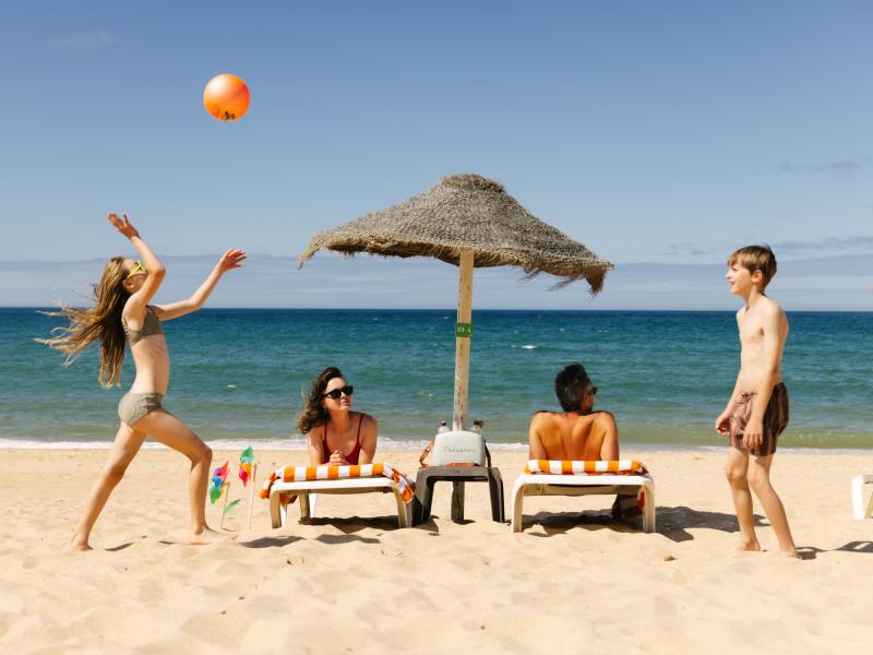 A family enjoys a day at the beach, with a girl playing with an orange ball and a boy standing near lounge chairs.