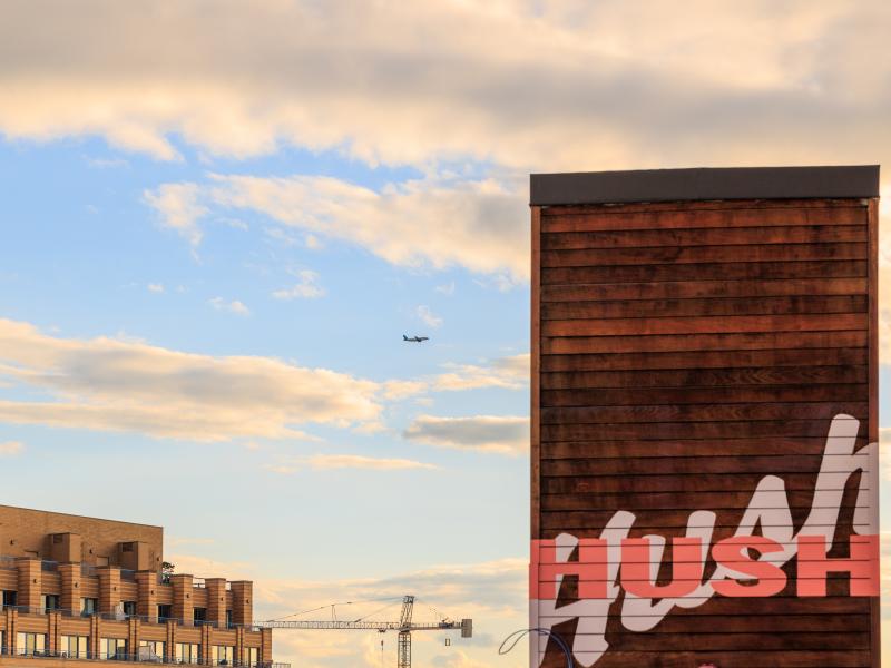 Wooden building facade with bold graffiti lettering, modern brick apartments and construction cranes beneath a partly cloudy sunset sky.