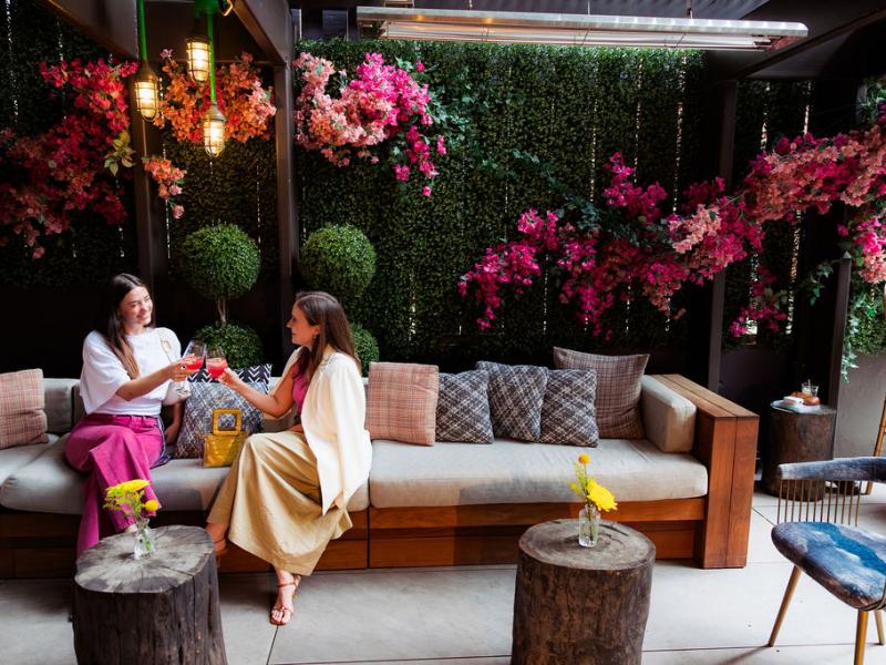Two women clink glasses while seated on a cushioned wooden sofa in a lush, flower-filled lounge.