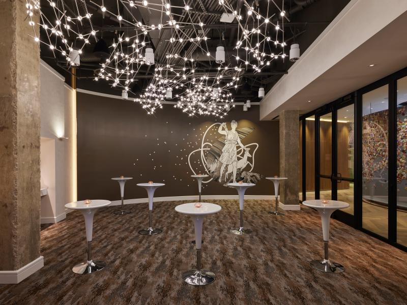 Um elegante salão para eventos com mesas de cocktail brancas, um mural decorativo na parede, portas de vidro e uma impressionante constelação de candeeiros suspensos no teto.