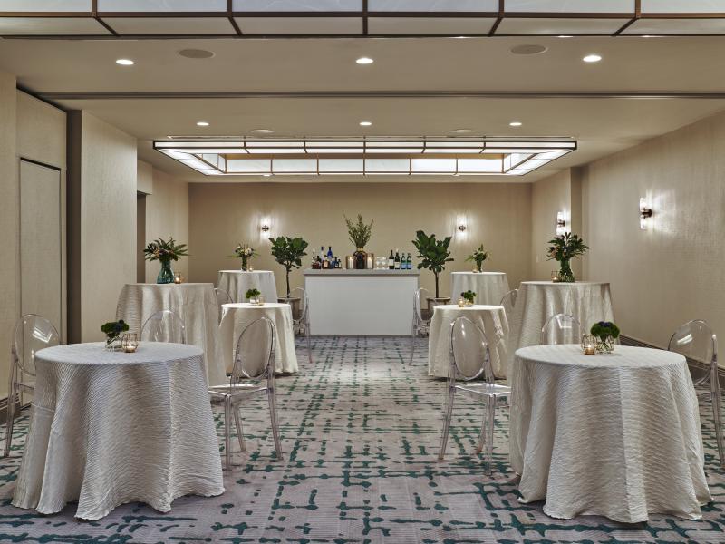 Elegant banquet room set for a reception with white-draped cocktail tables, floral centerpieces, clear chairs, and a stocked bar.