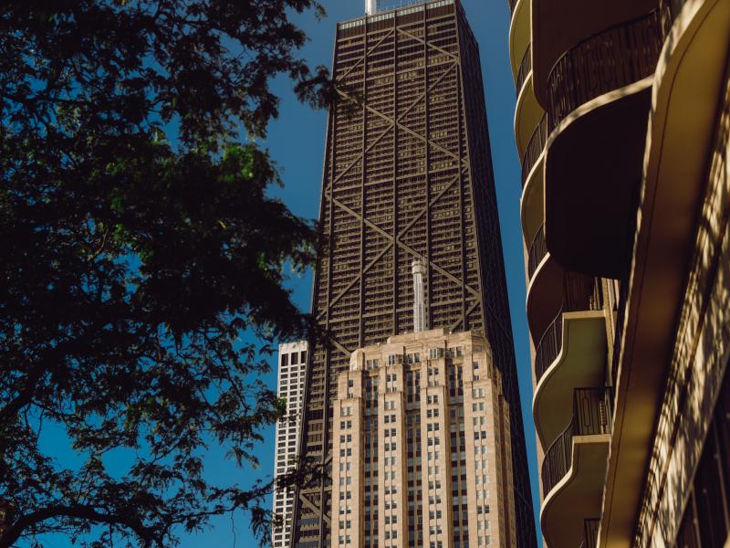 O John Hancock Center, um edifício escuro com fachadas em treliça, ergue-se entre árvores frondosas e um bloco de apartamentos curvo, com as suas antenas gémeas a destacarem-se contra o céu azul.